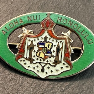 May include: A green oval-shaped enamel pin with a silver pin back. The pin features a white shield with a red, yellow, and blue design. The words "Aloha Nui Honolulu" are written in gold around the shield.