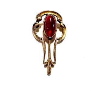 May include: A gold-toned metal brooch featuring a central oval-shaped deep red cabochon stone. The metal frame displays curved, symmetrical scrollwork details that extend downward into a tapered point, creating an elongated, ornate shape.