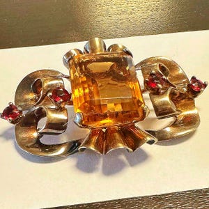 May include: A vintage gold-toned brooch featuring a large, rectangular amber-colored gemstone. The brooch has a ribbon-like design with three small, round, red gemstones. The brooch is a statement piece.