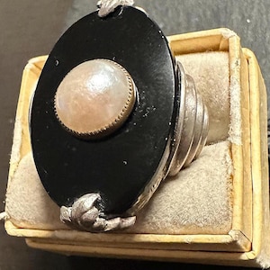 May include: An oval-shaped black pendant with a central, round, off-white pearl set in a gold-colored bezel. The pendant is attached to a silver-colored ring with a layered design. The jewelry is presented in a small, beige box with a soft, cream-colored interior.