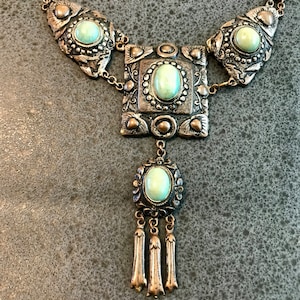 May include: A silver tone necklace with a square pendant featuring a turquoise stone and three dangling tassels. The necklace has a chain link design.