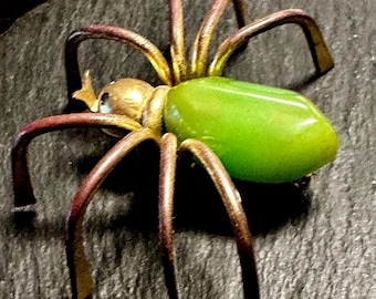 May include: A vintage spider brooch with a gold-tone body, brown legs, and a green oval-shaped abdomen. The brooch has eight curved legs and small blue accents on the head. The spider is a unique piece of jewelry.