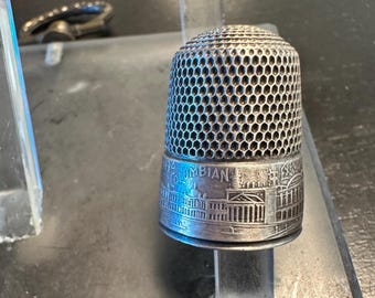 May include: A silver thimble with a honeycomb pattern on the top and a detailed architectural design around the base. The word "COLUMBIAN" is visible on the thimble.