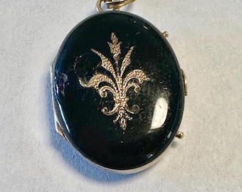 May include: Oval-shaped pendant with a dark green surface and a gold-colored fleur-de-lis design. The pendant has a gold-colored bail and a silver-colored frame. A classic piece of jewelry.