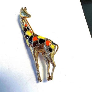 Puede incluir: Broche de jirafa dorada con manchas de esmalte de colores naranja, amarillo, marrón y negro. El broche es un diseño tridimensional detallado, que muestra el largo cuello y las patas de la jirafa. Un accesorio clásico con temática animal.