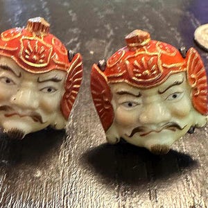 May include: Two antique Japanese ceramic figurines depicting a warrior's face with a stern expression. The figurines feature red and gold helmets, detailed facial features, and a small coin.