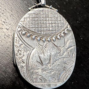 May include: Silver-toned oval locket with intricate engraved floral and sunburst designs. The locket features a decorative top with a bail for a chain. The metal has a vintage appearance.