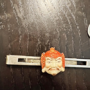 May include: A vintage tie clip featuring a detailed, orange and cream-colored face with a red helmet. The face has a mustache and beard. The clip is silver-toned and rectangular.