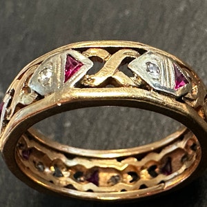 May include: A gold ring with a repeating pattern of alternating shapes. The ring features small, square red gemstones and tiny clear gemstones. The ring has a detailed, openwork design.