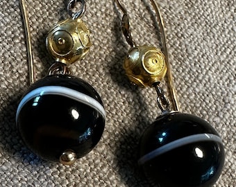 May include: A pair of gold earrings with black and white agate stone drops. The earrings have a gold ball charm above the stone drops.