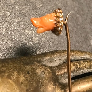May include: A vintage hat pin featuring a coral-colored, carved figure, possibly a bird's head, set with small pearls and gold-tone metal. The pin is displayed against a textured, gray background.