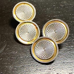 May include: Four vintage cufflinks with a silver and gold design. The cufflinks have a circular shape with a textured silver center and a gold-colored border. The background is a dark, textured surface.