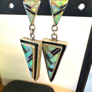 May include: A pair of dangle earrings featuring a triangular design. Each earring consists of two connected silver triangles inlaid with iridescent opal segments and thin black strips. The metal has a polished finish with visible dark oxidation.
