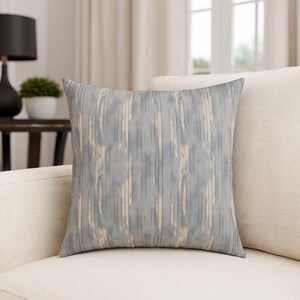 May include: Square decorative pillow with a vertical stripe pattern in shades of blue, gray, and beige. The pillow is placed on a cream-colored sofa. The pillow's design adds a touch of modern style to home decor.