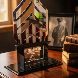 May include: A decorative military-themed award featuring a mirrored chevron design with red, white, and blue stripes and stars. The base includes text that reads "GT. DYLAN FOULKE" and a military emblem. A knife and crossed rifles are also included.