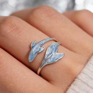 Può includere: Un anello d'argento con un design a delfino. Il corpo del delfino è azzurro chiaro con marmorizzazioni bianche. L'anello si avvolge attorno al dito, con la coda e la testa del delfino che formano le estremità.