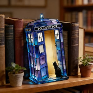 Puede incluir: Lámpara decorativa con forma de cabina telefónica azul, con paneles de vidrieras. La lámpara tiene las palabras "POLICE PUBLIC CALL BOX" en la parte superior. Una figura de gato negro está dentro. La lámpara está sobre una estantería de madera con libros.