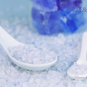 Puede incluir: Primer plano de cristales de sal azul persa en cucharas de cerámica blanca. Los cristales de sal son de color azul claro y están dispersos por la superficie. El texto "ROOT & RISE" es visible.