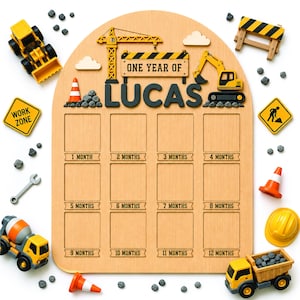 Pode incluir: Um quadro de marcos de madeira com topo arqueado, apresentando o texto "ONE YEAR OF LUCAS" e doze espaços retangulares para fotos rotulados de 1 mês a 12 meses. O quadro é decorado com recortes de construção, incluindo um guindaste, escavadeira e cones de trânsito, cercado por veículos de construção em miniatura e placas.
