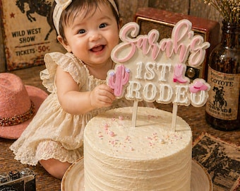 Könnte beinhalten: Ein Baby in einem cremefarbenen Kleid und Schleifen-Stirnband lächelt und hält einen Tortenaufsatz mit der Aufschrift "Sarah's 1st Rodeo". Der runde, glasierte Kuchen ist mit Streuseln verziert. Ein Vintage-Schild und eine Flasche sind im Hintergrund.