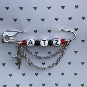 May include: A silver safety pin with the letters "ATZ" spelled out in white letter beads. The pin has red and black beads on either side of the letters, with a star charm and chain detail.