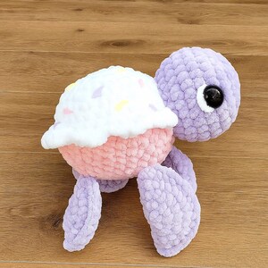 May include: A handmade crochet turtle plushie with a cupcake shell. The turtle has a lavender head, legs, and tail, a pink body, and a white top with colorful sprinkles. The turtle has a large black eye.