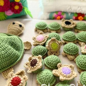 May include: Handmade crochet turtle toys in various stages of completion. The turtles feature green shells, tan bodies, and colorful flower accents. A large turtle body and head are also present, along with a green floral-decorated pouch.