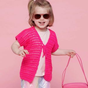 May include: A young child wearing a bright pink, open-front, short-sleeved crochet cardigan. The child is also wearing sunglasses and holding a pink purse. The background is a solid pink color.