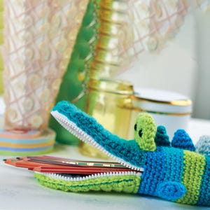 May include: Crocheted alligator pencil case in shades of blue and green, with a white zipper. The pencil case is open, revealing several colored pencils inside. The alligator has small, black eyes and a textured design.
