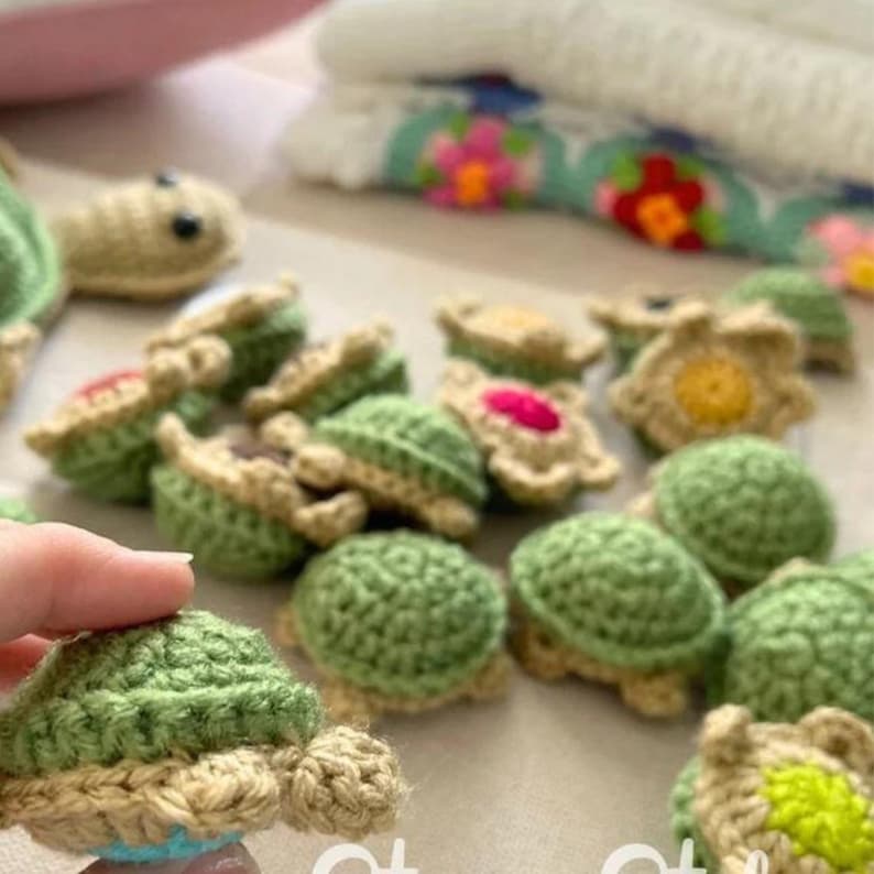 May include: A collection of handmade crochet turtles in various shades of green and tan. The turtles have detailed shells and are arranged in a group. Some turtles have pink or yellow accents. A hand is holding one of the turtles.
