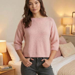 May include: A light pink sweater with three-quarter sleeves. The sweater has a boat neck and a ribbed texture. The person in the image is wearing dark gray jeans. The background includes a bed, nightstand, and lamps.