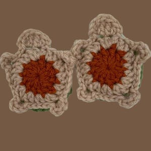 May include: Two handmade crochet turtle appliques. Each turtle has a tan body with a rust-colored center and green accents. The turtles are displayed on a brown background. These appliques are ideal for crafting projects.