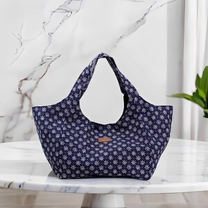 Peut inclure: Un sac fourre-tout bleu marine avec un motif floral blanc. Le sac a deux anses et une petite &eacute;tiquette rectangulaire en cuir marron. Le sac est pos&eacute; sur une table en marbre blanc.