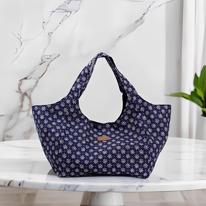 Peut inclure: Un sac fourre-tout bleu marine avec un motif floral blanc. Le sac a deux anses et une petite &eacute;tiquette rectangulaire en cuir marron. Le sac est pos&eacute; sur une table en marbre blanc.