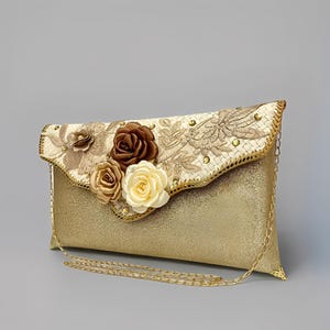 Könnte beinhalten: Goldfarbene Glitzer-Clutch mit dekorativem Überschlag, verziert mit floralen Stickereien und Stoffrosen in Braun- und Cremetönen. Die Clutch hat einen goldfarbenen Kettenriemen.