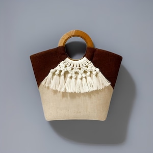 Puede incluir: Bolso tote con cuerpo beige natural y parte superior marrón oscuro. Cuenta con un asa de madera y un diseño de macramé blanco con borlas. El bolso está diseñado para transportar objetos.