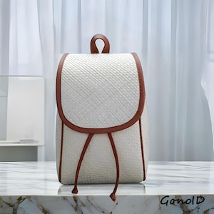 May include: A white woven backpack with brown trim and straps. The bag has a curved flap closure and a small handle at the top. The bag is sitting on a marble surface.