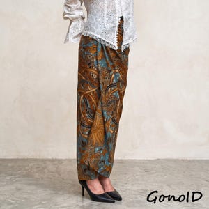 May include: A long, wrap-style skirt featuring a detailed brown and teal batik pattern with floral and leaf motifs. The skirt is paired with a white, long-sleeved blouse that has an eyelet lace design and scalloped edges along the cuffs and front.