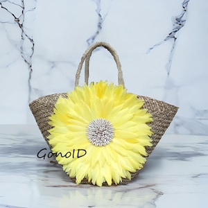 Puede incluir: Un bolso tote de paja tejida con una gran flor de plumas amarilla brillante en la parte delantera. La flor tiene un centro blanco y gris. El bolso tiene asas tejidas y el texto "GonoID" es visible.
