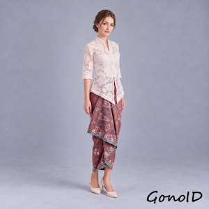 Könnte beinhalten: Eine cremefarbene Spitzenbluse mit Dreiviertelärmeln und Stehkragen, kombiniert mit einem langen, kastanienbraunen Batikrock mit komplexen geometrischen und floralen Mustern in Blau und Weiß. Das Outfit wird durch helle, spitze Pumps ergänzt.