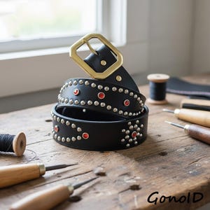 May include: A black leather belt with silver studs and red accents. The belt features a large gold-colored buckle. The belt is coiled on a wooden surface, with leatherworking tools and spools of thread in the background.