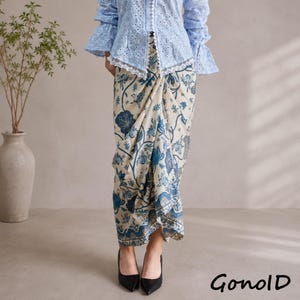 Pode incluir: Uma blusa azul clara de renda com ilhós e mangas compridas combinada com uma saia envelope creme com uma estampa floral e botânica azul. A saia tem um design drapeado na frente e cai até os tornozelos. O texto GonolD é visível no canto.