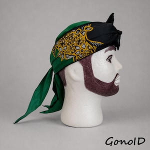 Könnte beinhalten: Eine grün-schwarze Kopfbedeckung mit einem detaillierten gold-weißen floralen Batikmuster. Der Stoff ist hinten mit zwei langen, spitz zulaufenden Enden gebunden. Die Kopfbedeckung wird auf einem weißen Mannequinkopf mit braunem Bart präsentiert.