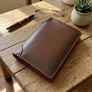 May include: A brown leather journal with red interior and white stitching sits on a wooden surface. A fountain pen and round eyeglasses are next to the journal. A small potted succulent is in the background.