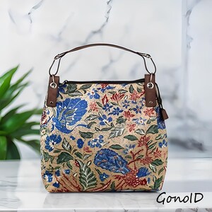 Pode incluir: Uma bolsa tote bege com um padrão floral em azul, vermelho e verde. A bolsa tem alças e detalhes em couro castanho. A bolsa tem formato quadrado e fecho de zíper. O texto "Gonold" é visível no canto inferior direito.