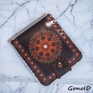 May include: A dark brown leather journal featuring a vibrant mandala design on the cover. The edges are stitched with brown thread, and the closure has a button. The journal has the text "GonolD" in the bottom right corner.