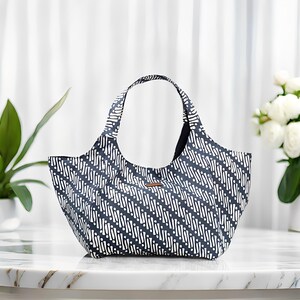 Peut inclure: Un sac fourre-tout avec un motif batik noir et blanc. Le sac a une forme arrondie avec une poign&eacute;e et une petite &eacute;tiquette marron. L'arri&egrave;re-plan comprend un rideau blanc, une plante et un vase de fleurs blanches.