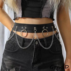 Może przedstawiać: Czarny pasek z metalowymi pierścieniami i łańcuchami w kolorze srebrnym. Pasek jest noszony na czarnych dżinsach. Pasek ma estetykę punk lub alternatywną.