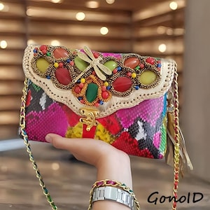 Può includere: Una pochette colorata con tracolla a catena. La borsa presenta un vivace motivo a pelle di serpente in rosa, rosso e giallo. La patta è decorata con pietre preziose e una libellula dorata. La borsa ha una chiusura dorata e la parola "Gonold" è stampata in basso a destra.