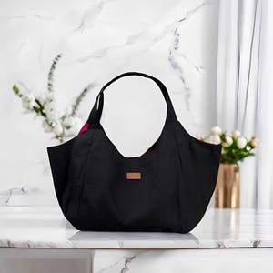 Peut inclure: Un sac fourre-tout noir avec un design large et ouvert et deux poign&eacute;es incurv&eacute;es. Le sac a une petite &eacute;tiquette marron rectangulaire sur le devant. La doublure int&eacute;rieure est un tissu rose et rouge &agrave; motifs. Le sac est pos&eacute; sur une surface en marbre blanc.